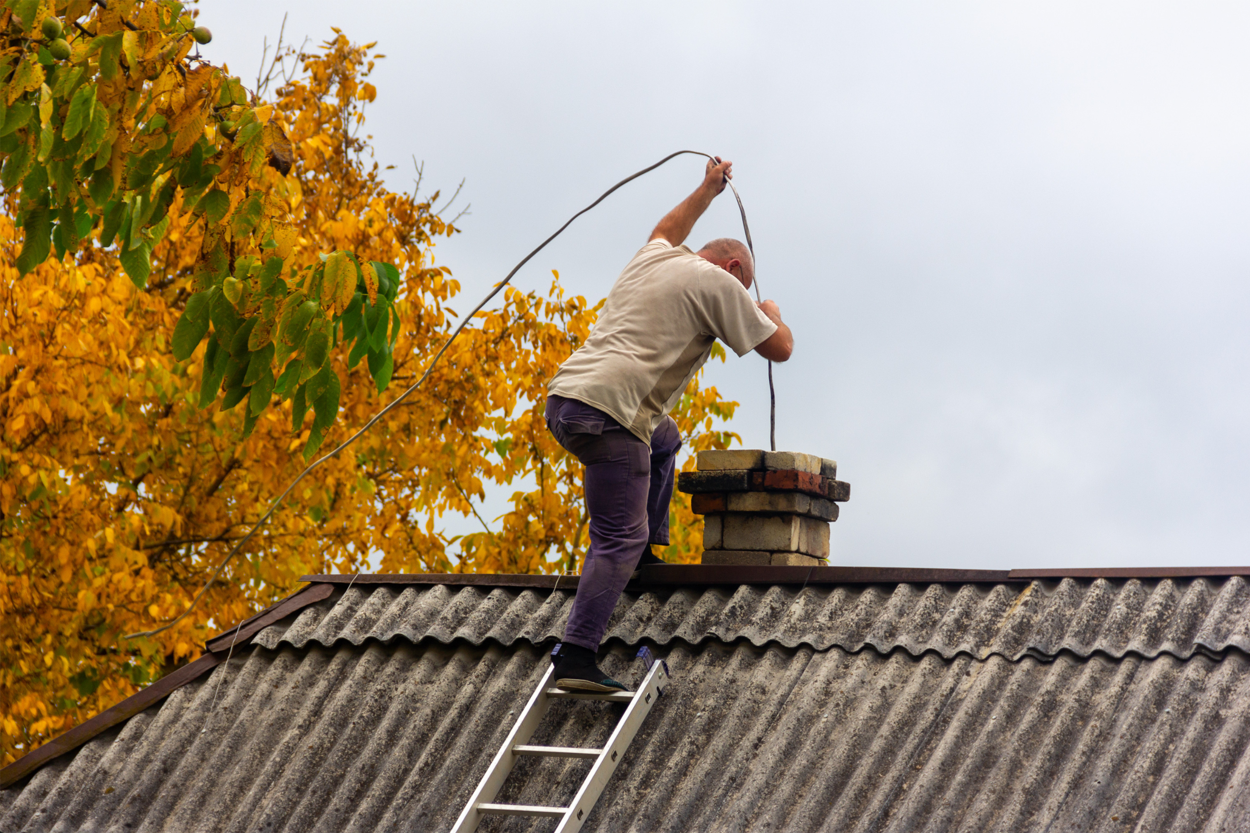 Homme de dos réalisant le ramonage préventif d'une cheminée depuis le toit de la maison